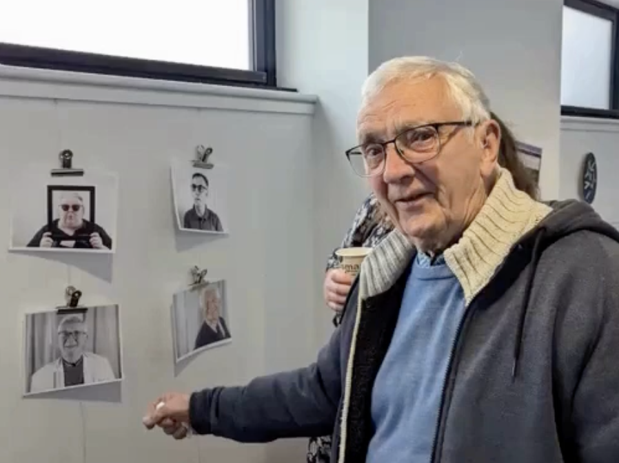 A man with silver hair and glasses points to his image on the wall of the gallery