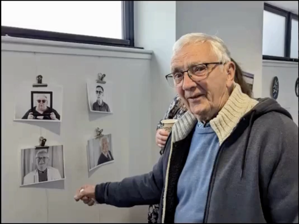 A man with silver hair and glasses points to his image on the wall of the gallery